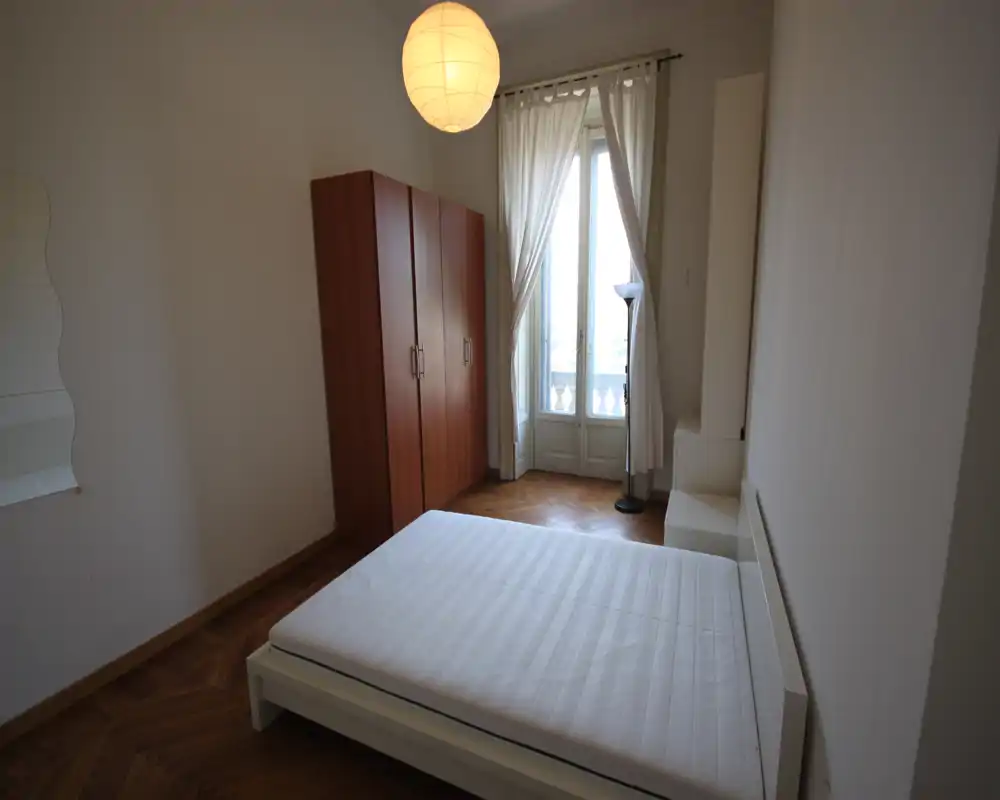 Dormitorio luminoso con cama doble en estructura blanca baja, suelo de parquet y ventana alta estilo francés con cortinas vaporosas que dejan pasar la luz natural. Armario sencillo y lámpara de pie completan el espacio minimalista.