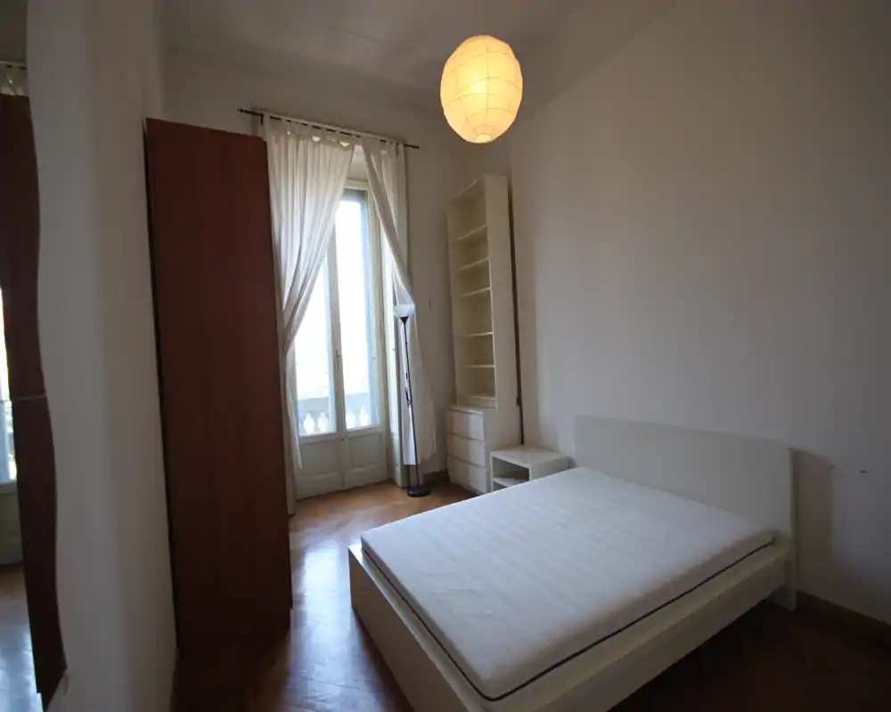 Dormitorio luminoso y minimalista con cama doble, ventana alta con cortinas translúcidas, estantería y suelo de parquet — espacio limpio y con mucha luz natural.