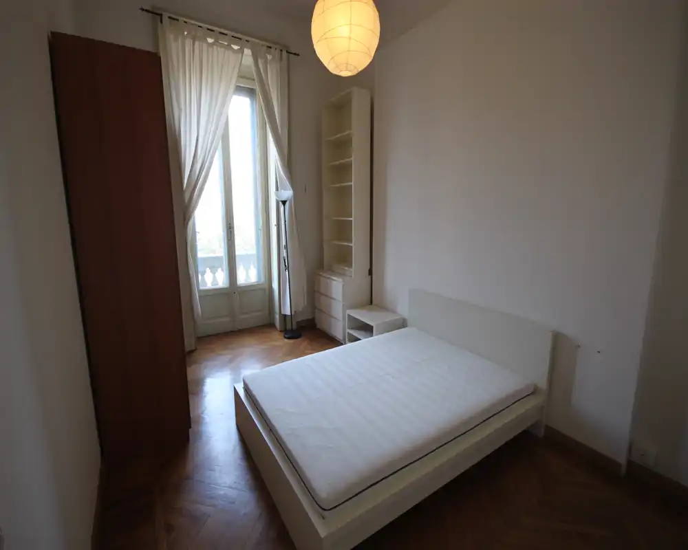 Dormitorio luminoso con cama doble, suelo de parquet y puerta balcón que aporta luz natural. Mobiliario blanco y sencillo con cabecero, mesita y estantería, ideal para un estilo limpio y minimalista.