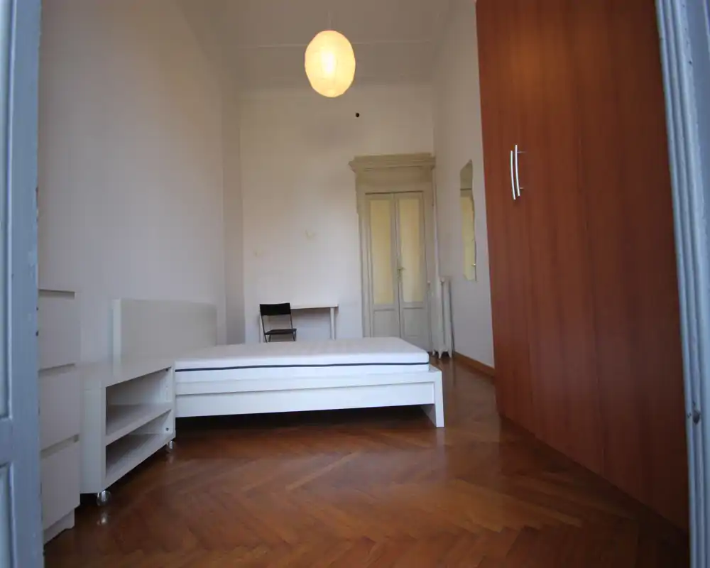 Amplio dormitorio con cama blanca centrada sobre suelo de parquet en espiga, armario a la derecha y lámpara colgante — luminoso y con mobiliario sencillo.