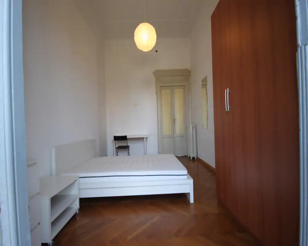 Dormitorio luminoso y sencillo con cama de estructura blanca, colchón, mesita y un gran armario de madera; techos altos y suelo de parquet que crean un espacio aireado y minimalista.