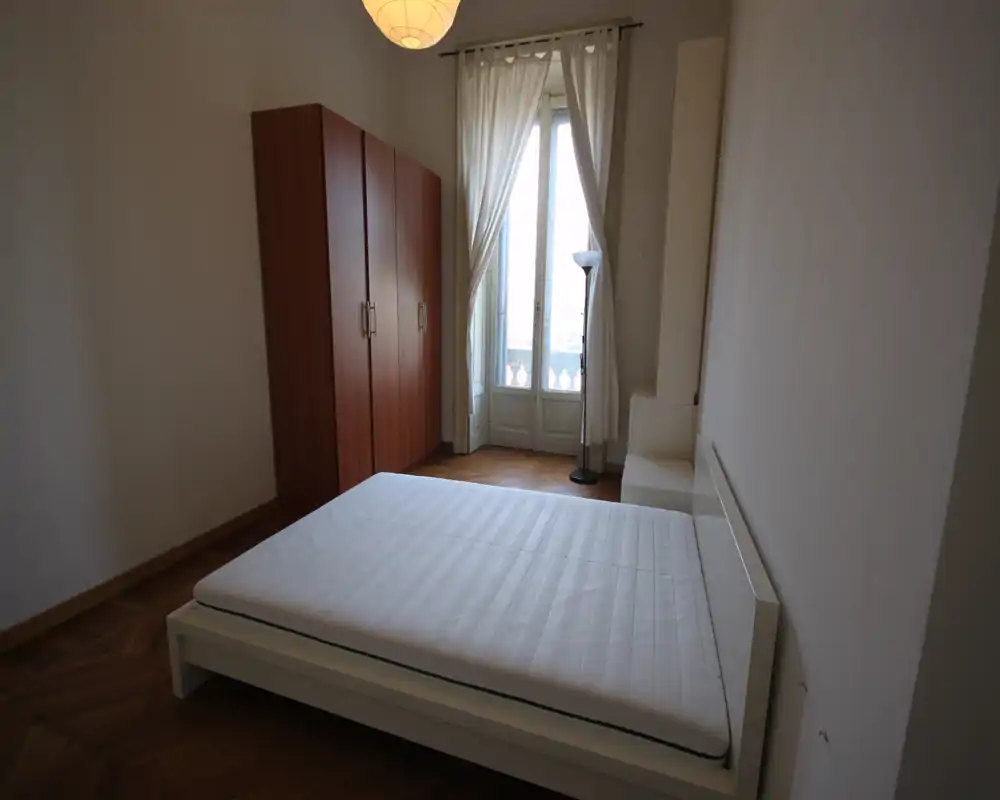 Dormitorio luminoso y minimalista con cama doble blanca, armario de madera y ventana alta que aporta luz natural. Líneas sencillas y suelo de parquet crean un ambiente tranquilo y aireado.