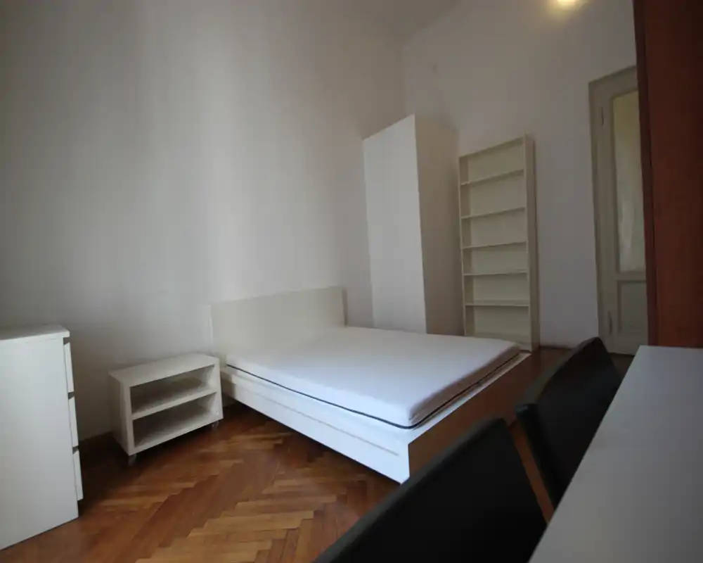 Dormitorio luminoso y de mobiliario sencillo con cama doble, mesita, armario y estantería. Estilo limpio, suelo de parquet y paredes neutras crean un espacio para dormir funcional y tranquilo.