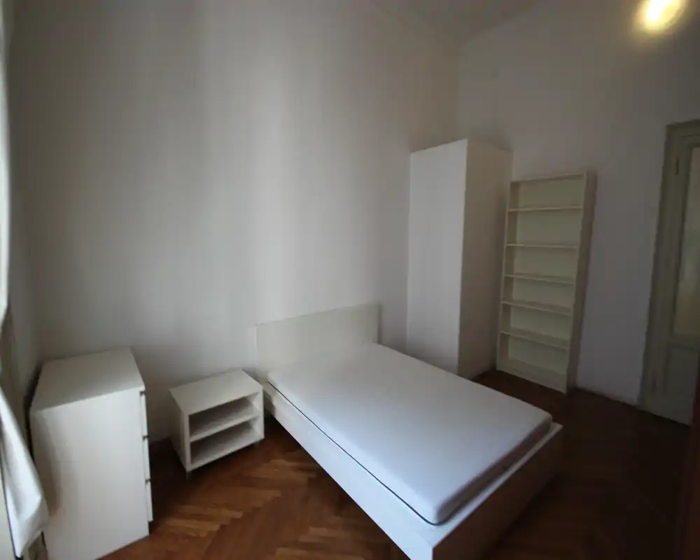 Dormitorio minimalista y luminoso con cama doble, mobiliario blanco y suelo de parquet. Distribución sencilla con armario, estanterías y mesita; apropiado para anuncios que destaquen decoración limpia y neutra.