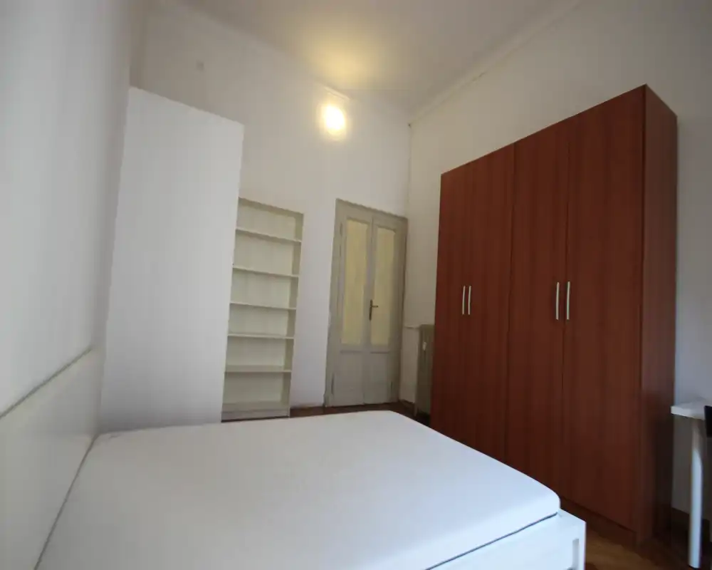 Dormitorio luminoso y sencillo con un gran colchón en primer plano, un armario de madera, estanterías y paredes blancas — espacio para dormir limpio y funcional.