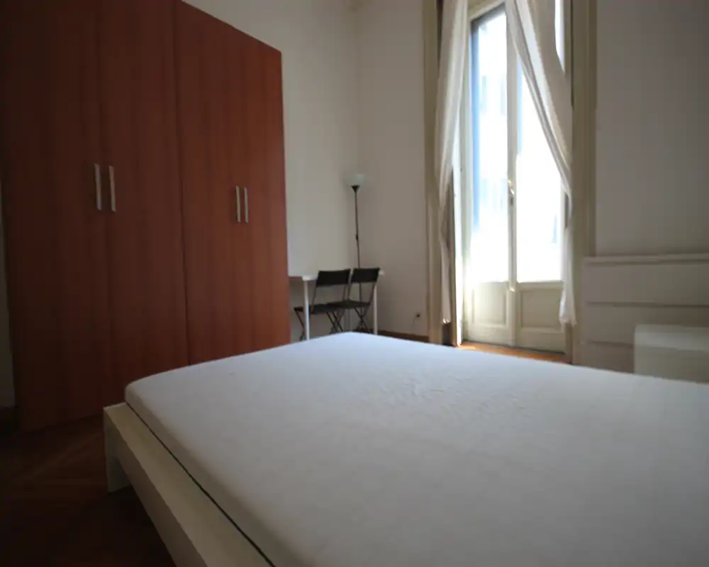 Dormitorio luminoso y amueblado de forma sencilla con un gran colchón en primer plano, un armario de madera, un pequeño escritorio y una ventana alta que aporta luz natural.
