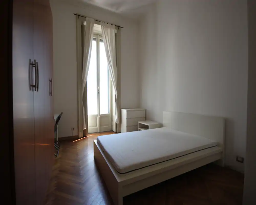 Dormitorio luminoso y minimalista con cama doble en estructura blanca baja, suelo de parquet y ventana alta con cortinas ligeras que dejan pasar la luz natural.