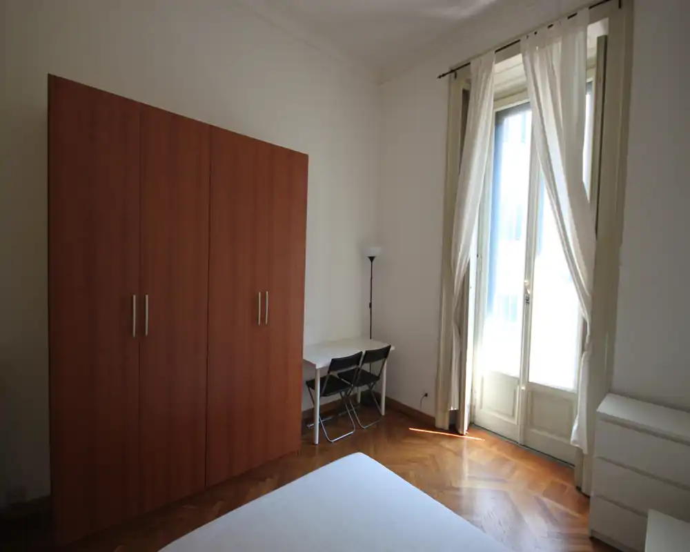 Dormitorio luminoso y sencillo con cama doble en primer plano, gran armario de madera, escritorio con dos sillas y ventana alta con cortinas blancas que dejan entrar luz natural.