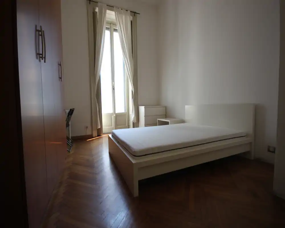 Dormitorio luminoso y minimalista con cama doble en estructura baja blanca, suelo de parquet y una gran ventana con cortinas que deja entrar luz natural.