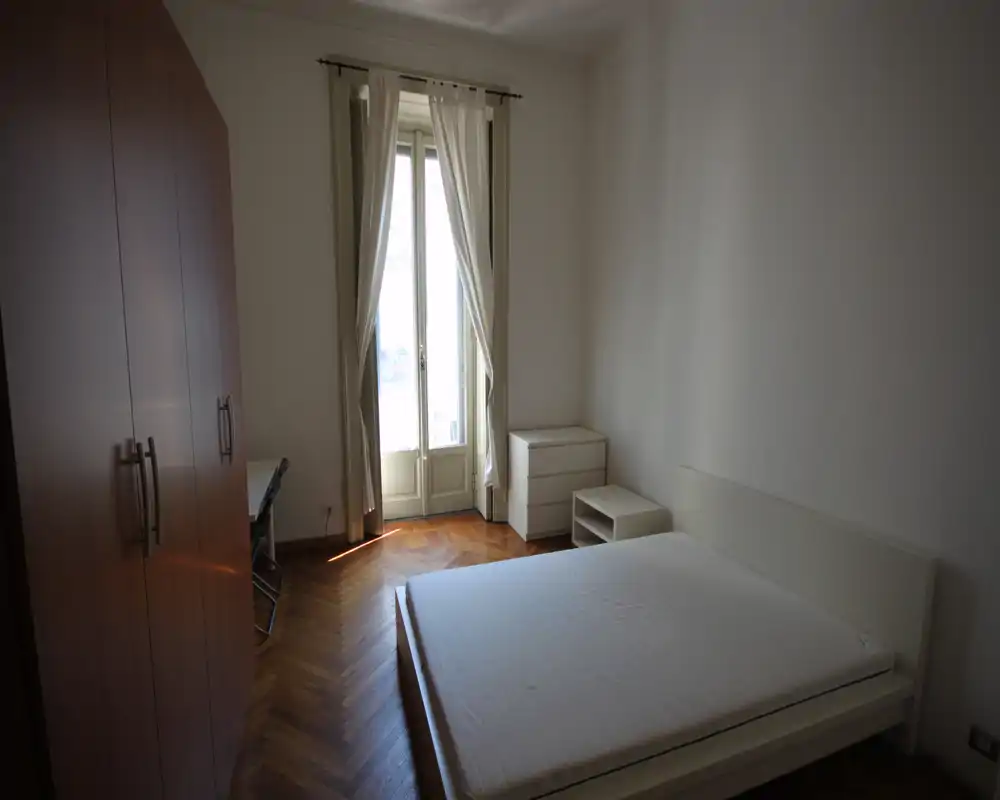 Dormitorio luminoso con cama doble, mobiliario blanco y ventana estilo francés que aporta luz natural. Suelo de parquet y armario ofrecen almacenamiento en una distribución limpia y sencilla.
