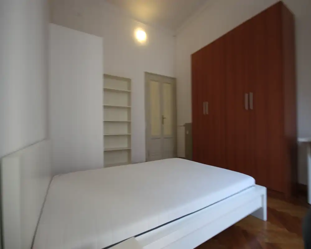 Dormitorio luminoso y sencillo con una cama doble blanca en primer plano, armario de madera y estanterías, suelo de parquet y paredes neutras: un espacio para dormir limpio y minimalista.