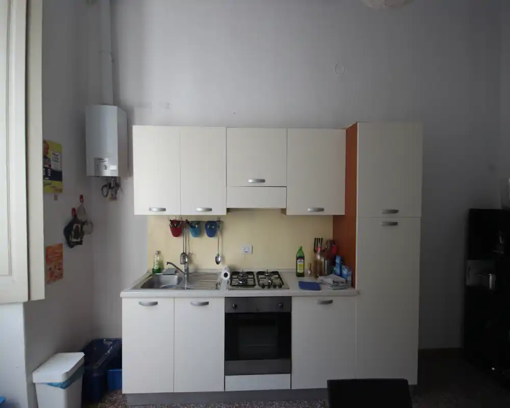 Cocina compacta equipada con muebles blancos, fregadero de acero y placa de cuatro fuegos; distribución ordenada y funcional con luz natural desde la ventana izquierda.