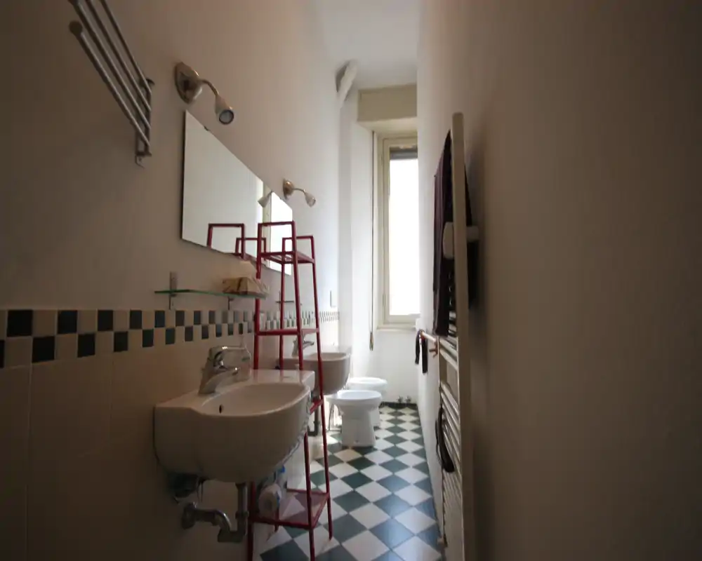 Baño estrecho y bien iluminado con dos lavabos de pared, bidé y váter, suelo de baldosas en patrón y una ventana alta que aporta luz natural.