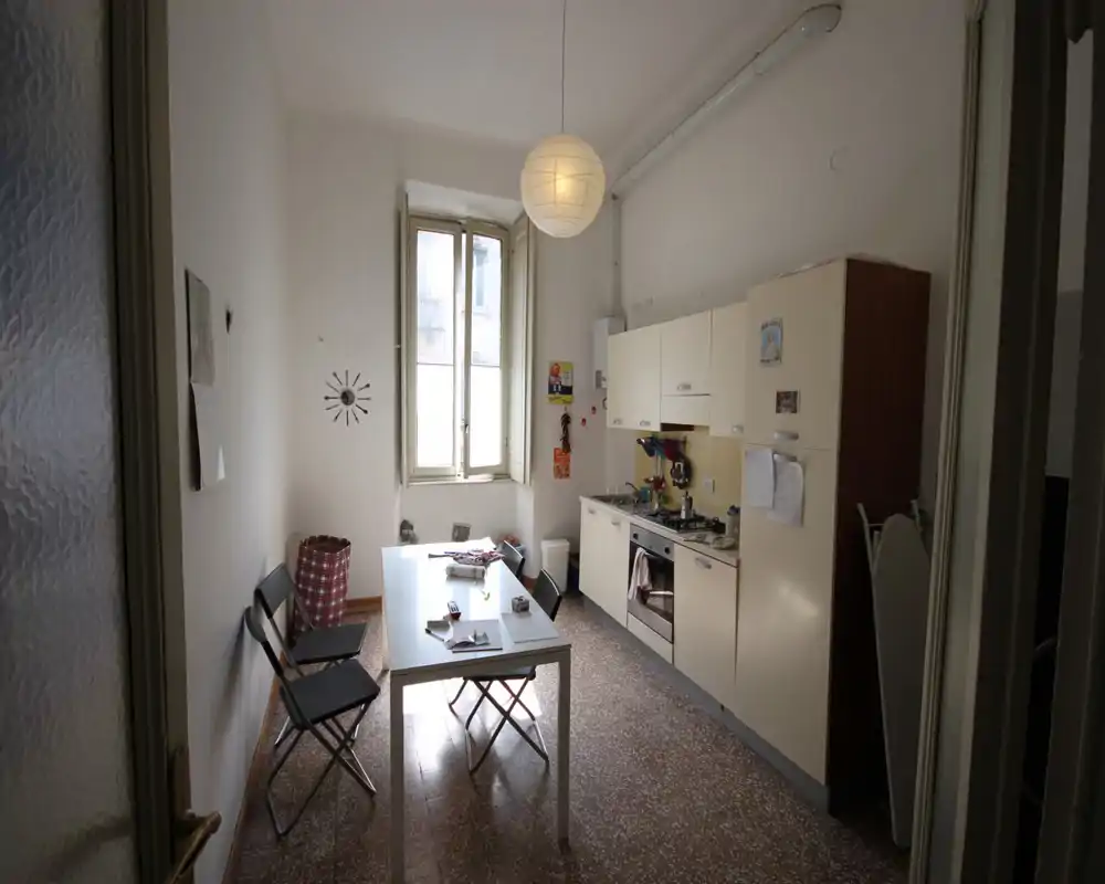 Cocina funcional con muebles crema, horno empotrado y una pequeña mesa de comedor bajo una gran ventana. El espacio está iluminado de forma natural y muestra objetos cotidianos que transmiten un ambiente práctico y habitado.