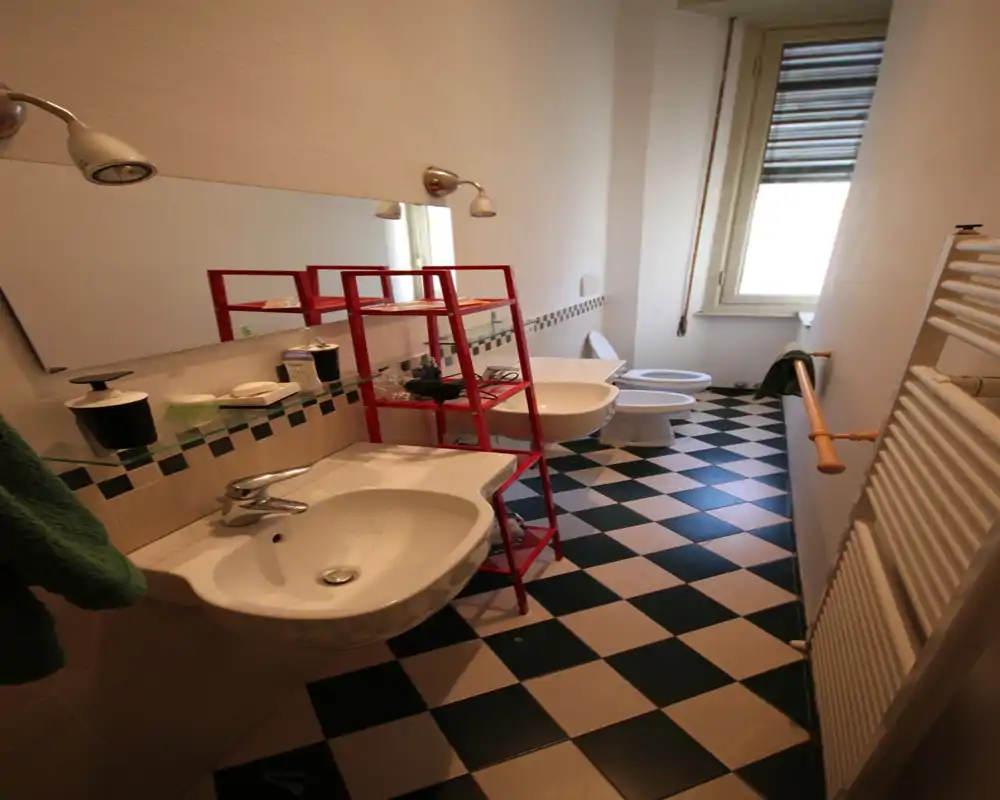 Baño estrecho con lavabo mural, bidé y inodoro, suelo ajedrezado y una ventana alta que aporta luz natural.