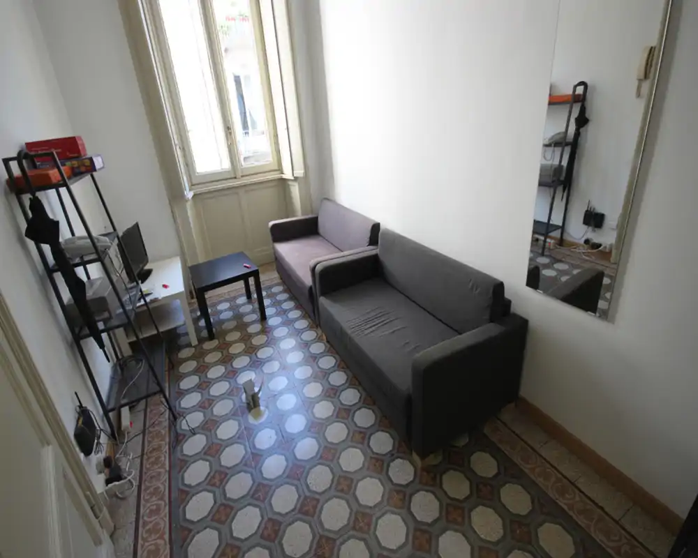 Salón compacto con dos sofás, una mesa de centro pequeña y una estantería alta; luz de ventana y suelos con baldosas decorativas que aportan carácter.