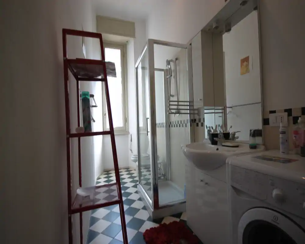 Baño compacto con cabina de ducha de vidrio, lavabo con mueble espejo y lavadora de carga frontal; suelo de baldosas en blanco y negro y luz natural desde una ventana alta.
