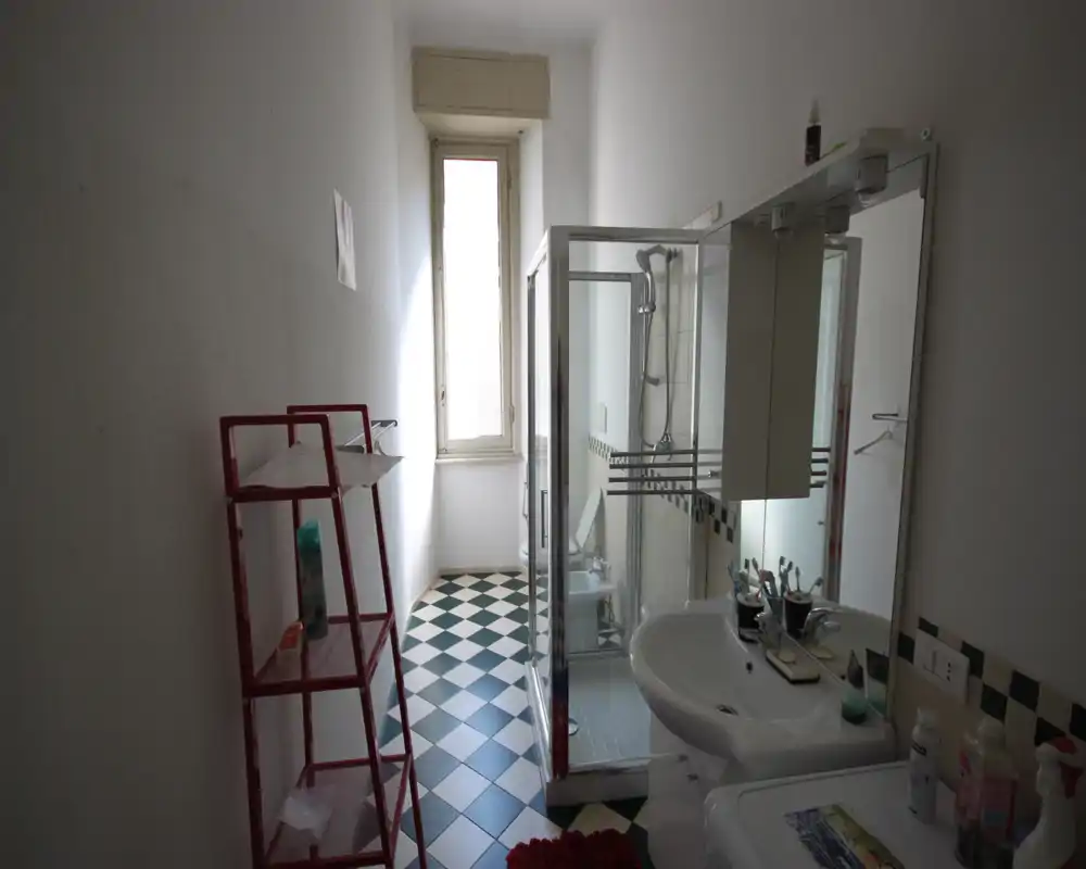 Baño compacto con cabina de ducha en esquina, lavabo con armario con espejo y una ventana alta y estrecha que aporta luz natural.