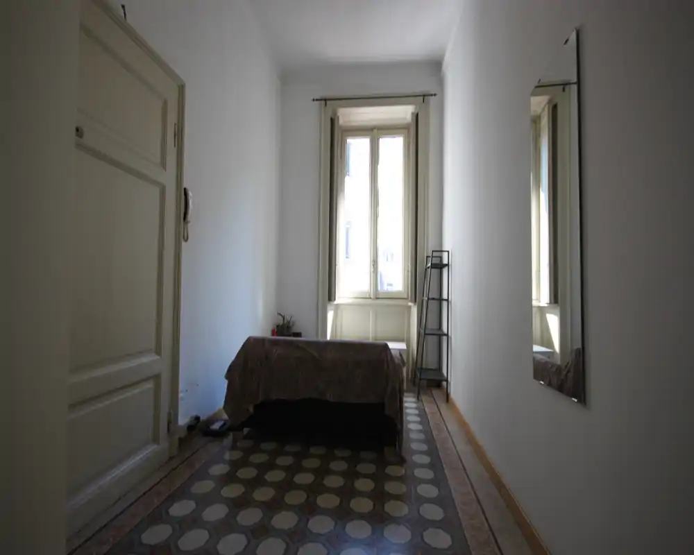 Dormitorio sencillo con cama individual junto a la ventana, techo alto y suelo de baldosas decorativas; luz natural entra por una ventana alta.