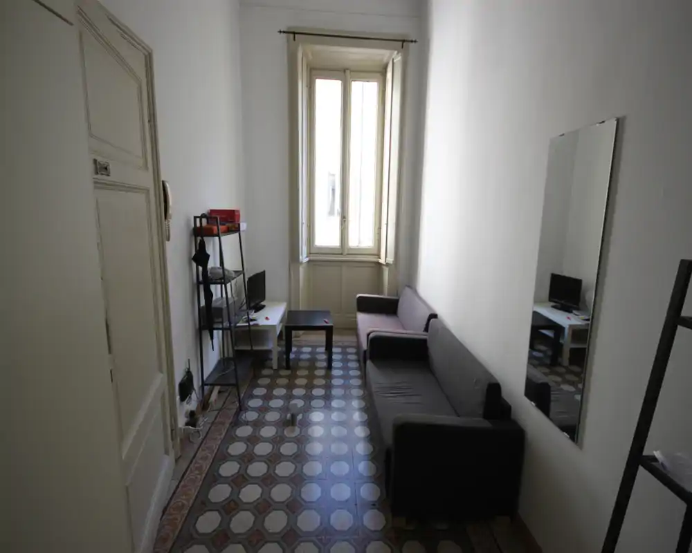 Salón estrecho con dos sofás, una mesa de centro pequeña y una ventana que aporta luz natural; el suelo hidráulico le da personalidad. Espacio funcional y minimalista, ideal para un piso compacto.