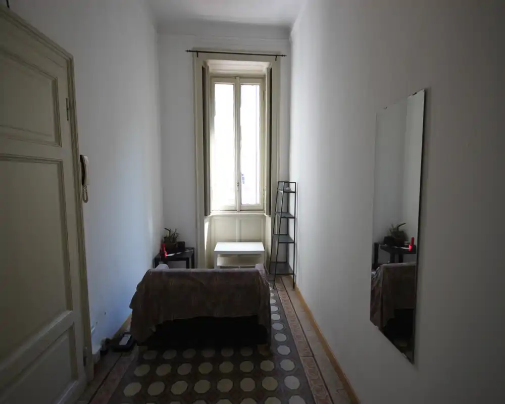Pequeña habitación con una cama/sofá individual junto a la pared bajo una ventana alta; mobiliario mínimo con mesita, estantería y espejo de cuerpo entero. Luz natural y suelos con baldosas decorativas le dan carácter.