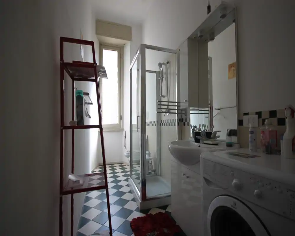 Baño luminoso y ordenado con cabina de ducha en esquina, lavabo con armario con espejo, lavadora y suelo de baldosas con patrón.