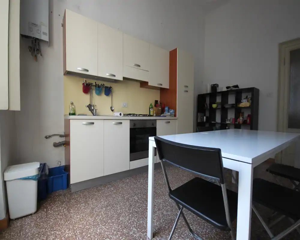 Cocina compacta y funcional con horno empotrado, armarios altos y una mesa blanca con sillas plegables — espacio luminoso y práctico para el uso diario.