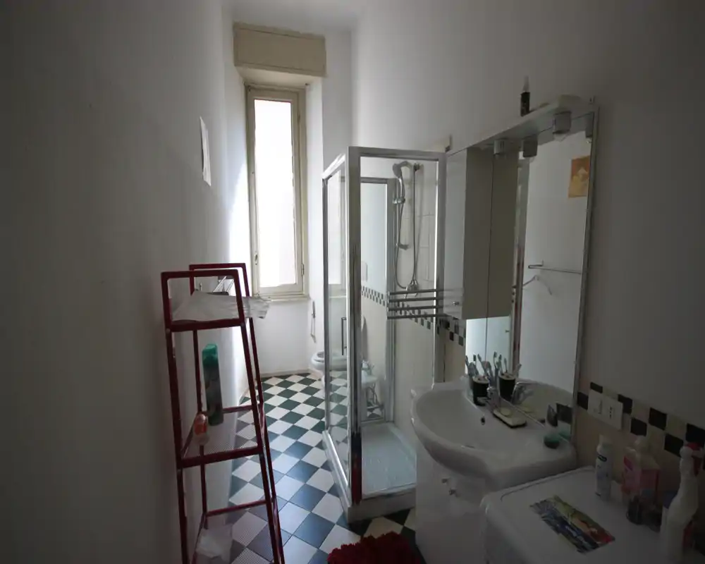 Baño luminoso y estrecho con mampara de ducha de cristal, lavabo, espejo con armario y suelo de baldosas decoradas; luz natural desde una ventana alta al fondo.
