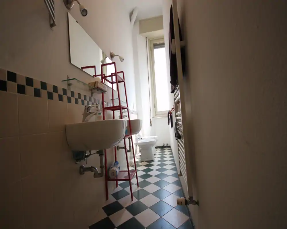 Baño estrecho con suelo a cuadros, dos lavabos suspendidos, inodoro al fondo y una ventana alta que aporta luz natural.
