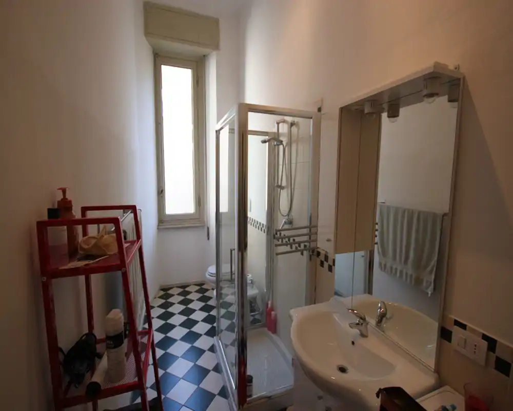 Baño estrecho y luminoso con cabina de ducha en esquina, lavabo blanco y suelo de baldosas ajedrezadas. Espacio limpio y funcional, ideal para un apartamento pequeño.