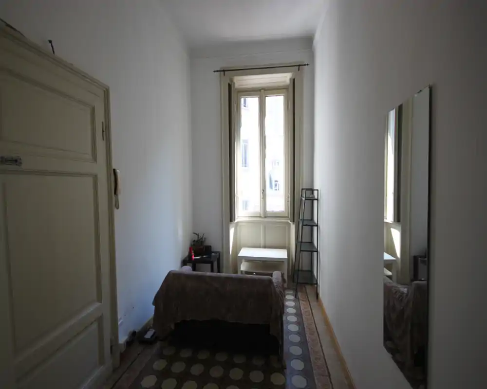 Habitación estrecha y luminosa con cama individual en el eje central, ventana alta que aporta luz natural, mesita y espejo en la pared; mobiliario mínimo y ordenado, ideal para una persona.