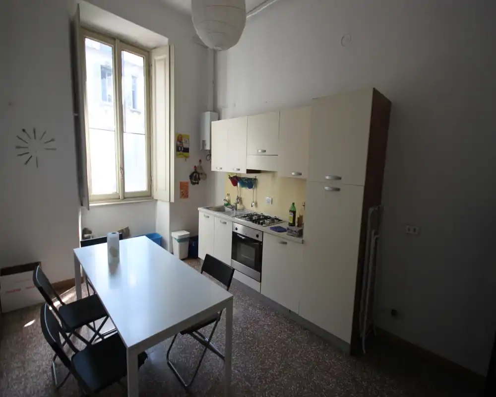 Cocina compacta y luminosa con una pared de muebles, horno empotrado y placa de gas, y una mesa con sillas plegables junto a una ventana alta.