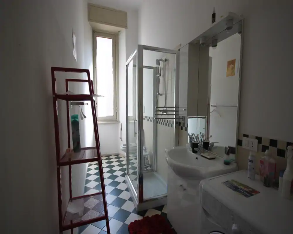Baño luminoso y ordenado con cabina de ducha de cristal, lavabo con mueble espejo, lavadora y suelo de baldosas con patrón.