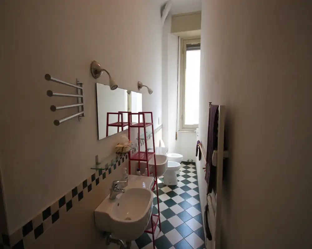 Baño estrecho y luminoso con dos lavabos suspendidos, inodoro y bidé al fondo, suelo de baldosas de patrón y una ventana alta que aporta luz natural.