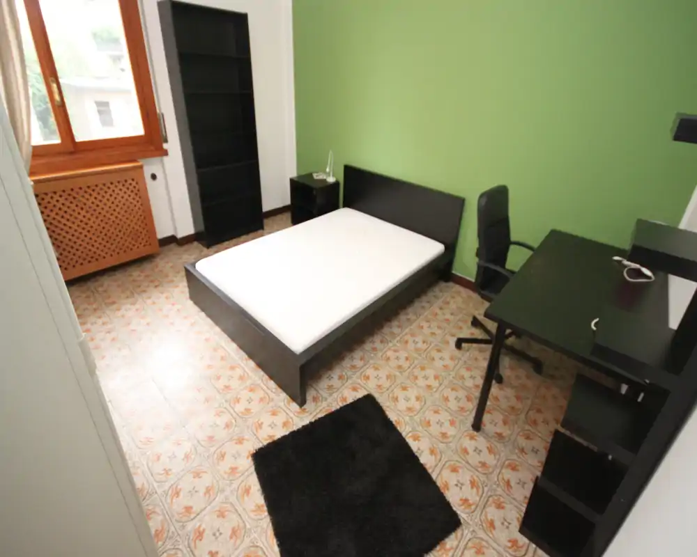 Chambre lumineuse et sobrement meublée avec un lit bas, un bureau et des étagères. La lumière naturelle venant de la fenêtre et les lignes nettes en font un espace pratique, idéal pour étudiants ou locations courtes.