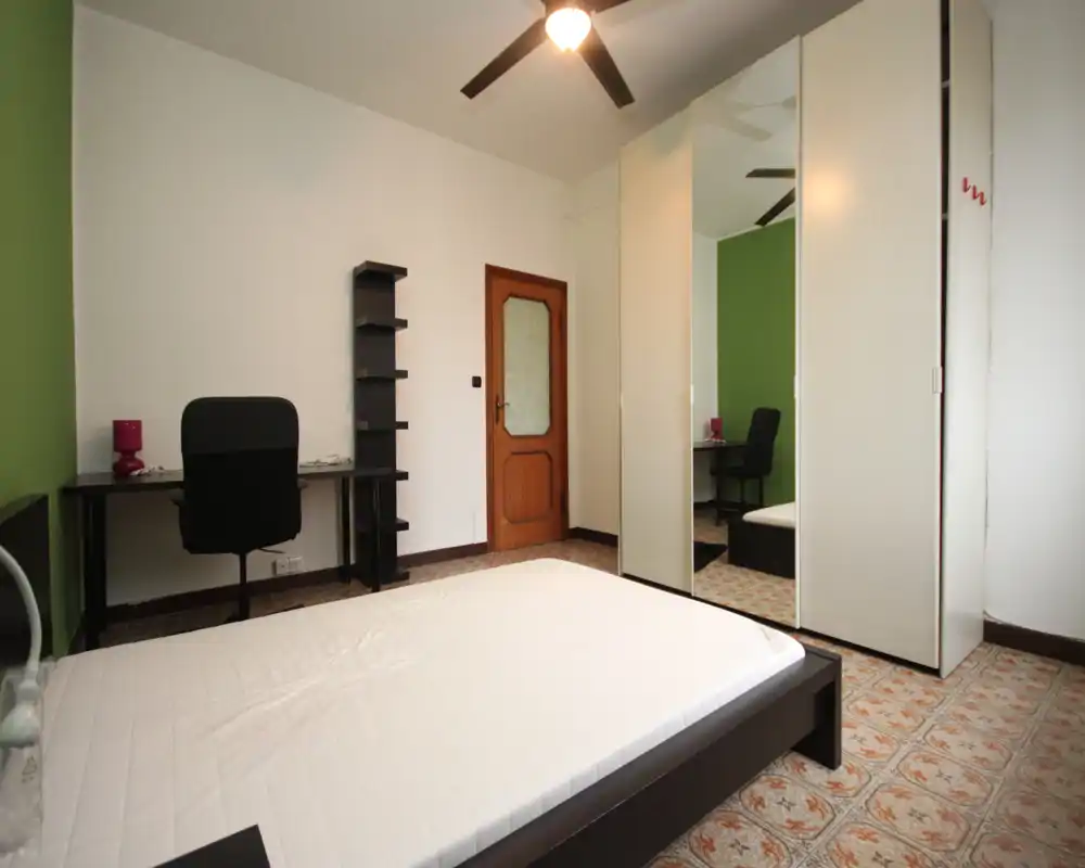 Chambre lumineuse et bien rangée avec un lit double au premier plan, une armoire miroir et un petit bureau contre le mur — simple, fonctionnelle et bien éclairée.