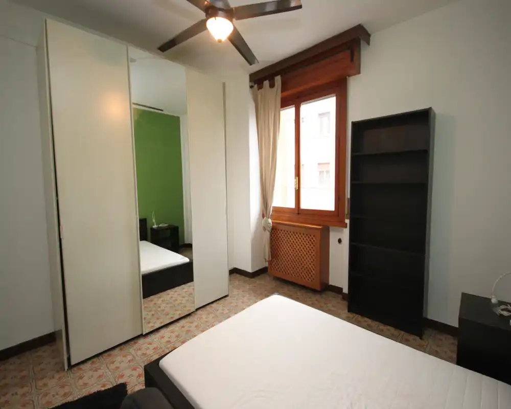 Chambre lumineuse et sobre avec un lit double, une armoire à portes miroir et une grande fenêtre laissant entrer la lumière naturelle. Sol carrelé à motif, ventilateur de plafond et mobilier fonctionnel.