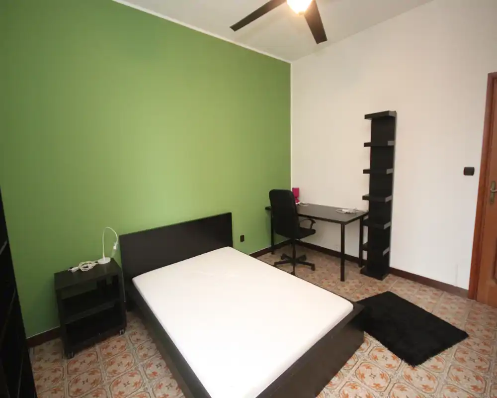 Chambre lumineuse et sobrement meublée avec grand lit, mur vert accentué, table de chevet et bureau avec étagère — idéale pour une personne ou un couple. Sol et éclairage ambiant bien visibles.
