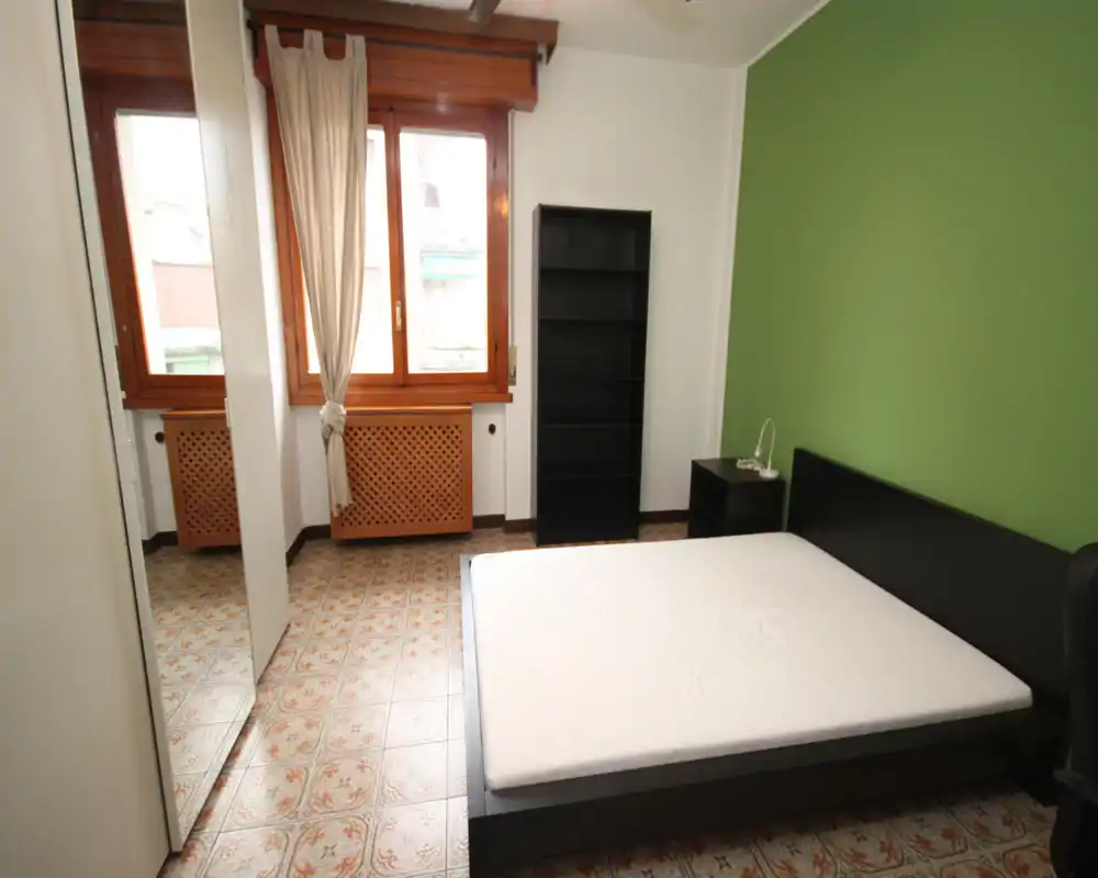 Chambre lumineuse avec un lit bas en bois foncé et matelas blanc, mur d'accent vert ; grande fenêtre apportant la lumière naturelle et une étagère sans portes pour le rangement.