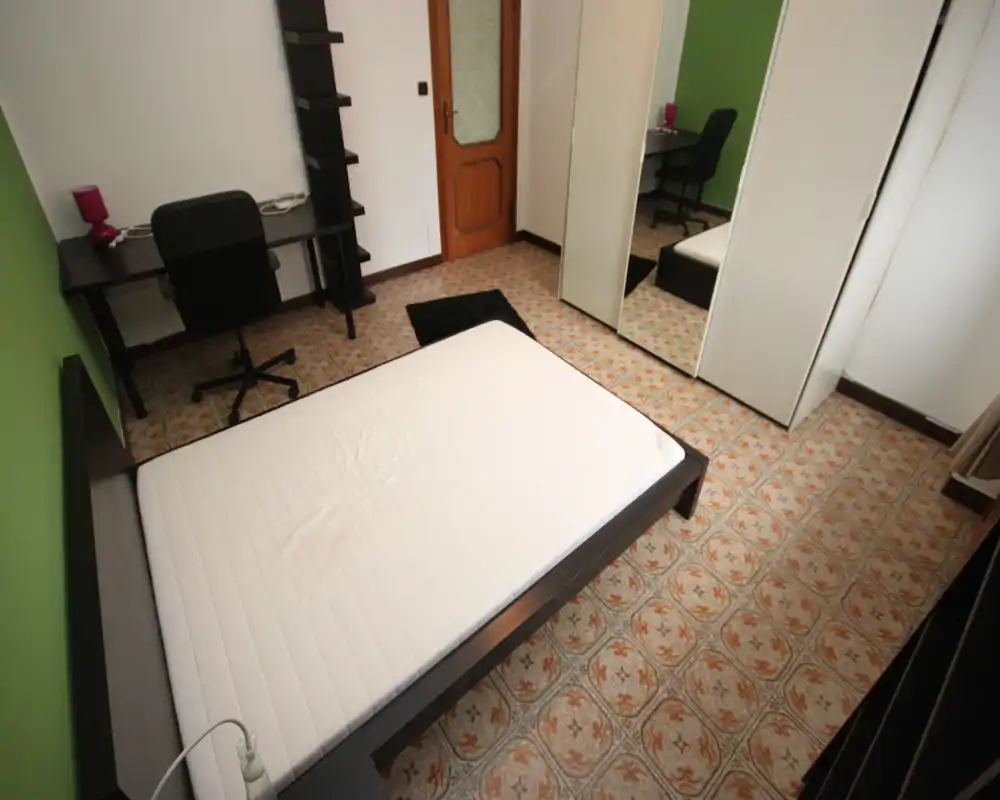 Chambre lumineuse et sobrement meublée avec un lit double sur cadre bas foncé, une armoire miroir, un bureau et une chaise — adaptée pour une personne ou un couple. Sol en carreaux à motif et mur accent vert.