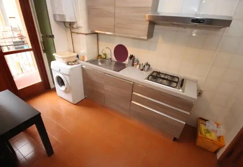 Cocina compacta y luminosa con muebles de madera, fregadero de acero, placa de gas y lavadora junto a la puerta del balcón — distribución práctica para un piso pequeño.