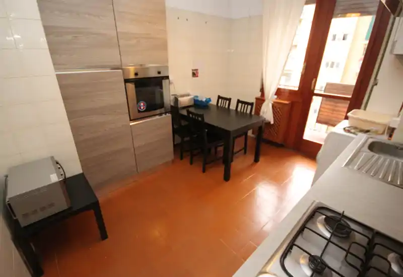 Cocina luminosa y práctica con horno empotrado, vitrocerámica de gas y mesa para cuatro junto a la puerta del balcón — ordenada y lista para usar.