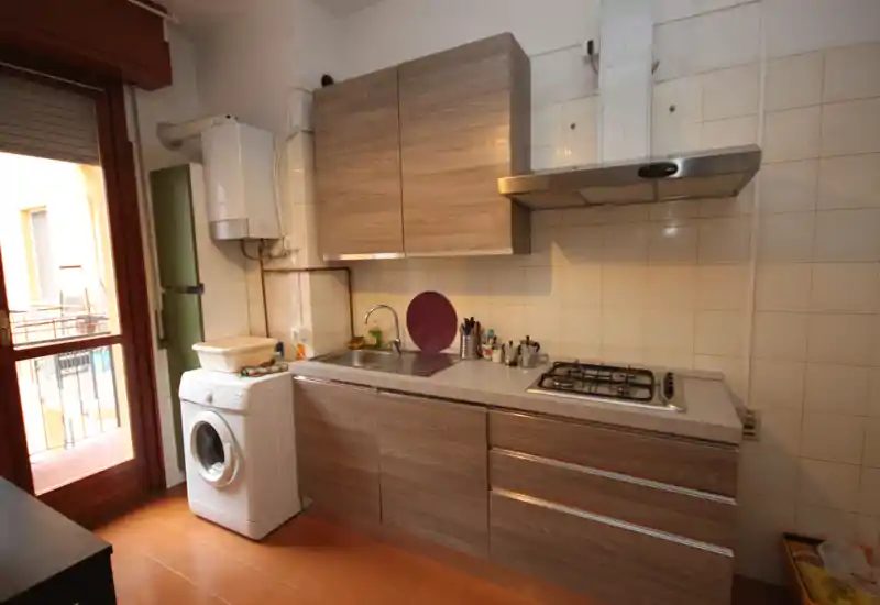 Cocina compacta y bien organizada con muebles efecto madera, fregadero de acero, placa de gas y una lavadora junto a la puerta del balcón que aporta luz natural.