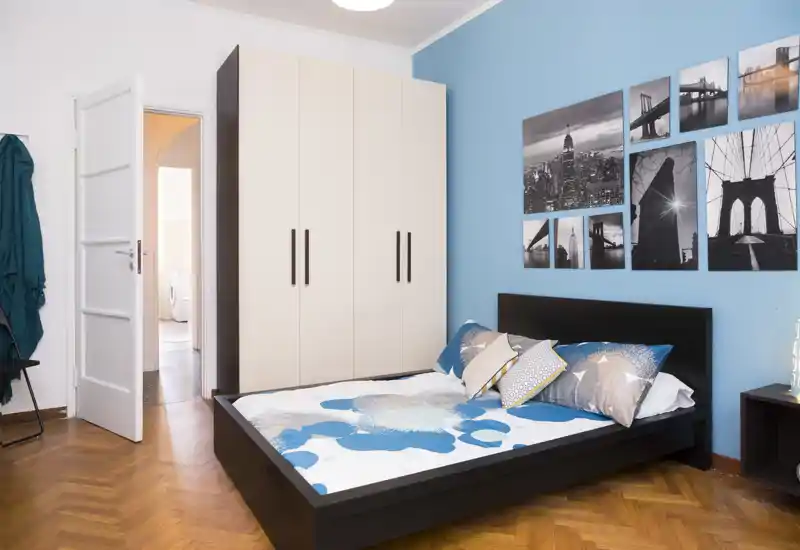 Camera da letto luminosa e curata con letto matrimoniale contro una parete blu, cuscini coordinati e un grande armadio; parquet in legno e foto in bianco e nero completano l'arredo.
