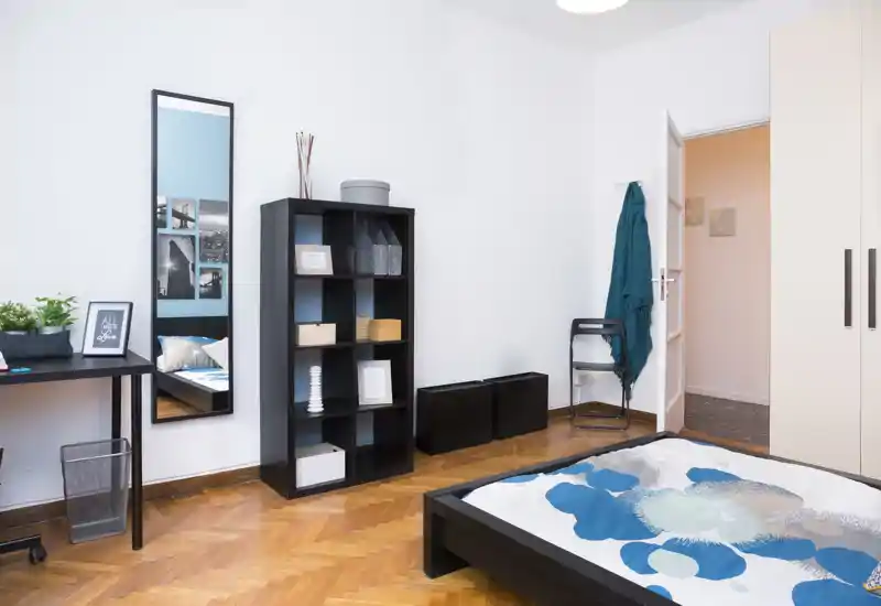 Camera da letto luminosa e moderna con un letto basso parzialmente visibile in primo piano, una libreria nera, uno specchio a figura intera e una scrivania: stile minimal pulito e parquet caldo.