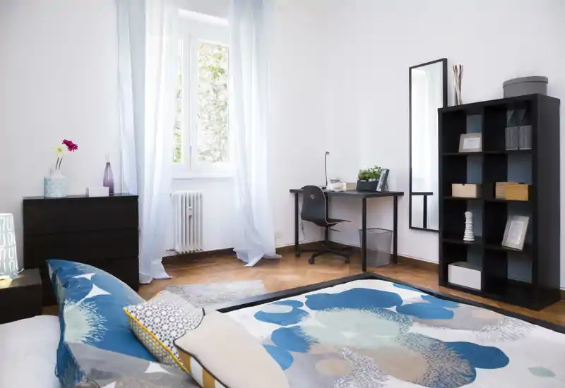Camera da letto luminosa e moderna con copriletto decorato, cuscini e una comoda postazione di lavoro vicino alla finestra. Luce naturale, parquet in legno e scaffalatura elegante creano un ambiente fresco e accogliente, perfetto per un annuncio.