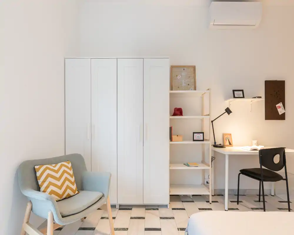 Bright, minimal bedroom corner with a wardrobe, shelving and a small desk. The space features a cozy armchair with a patterned cushion and warm task lighting ideal for reading or working.