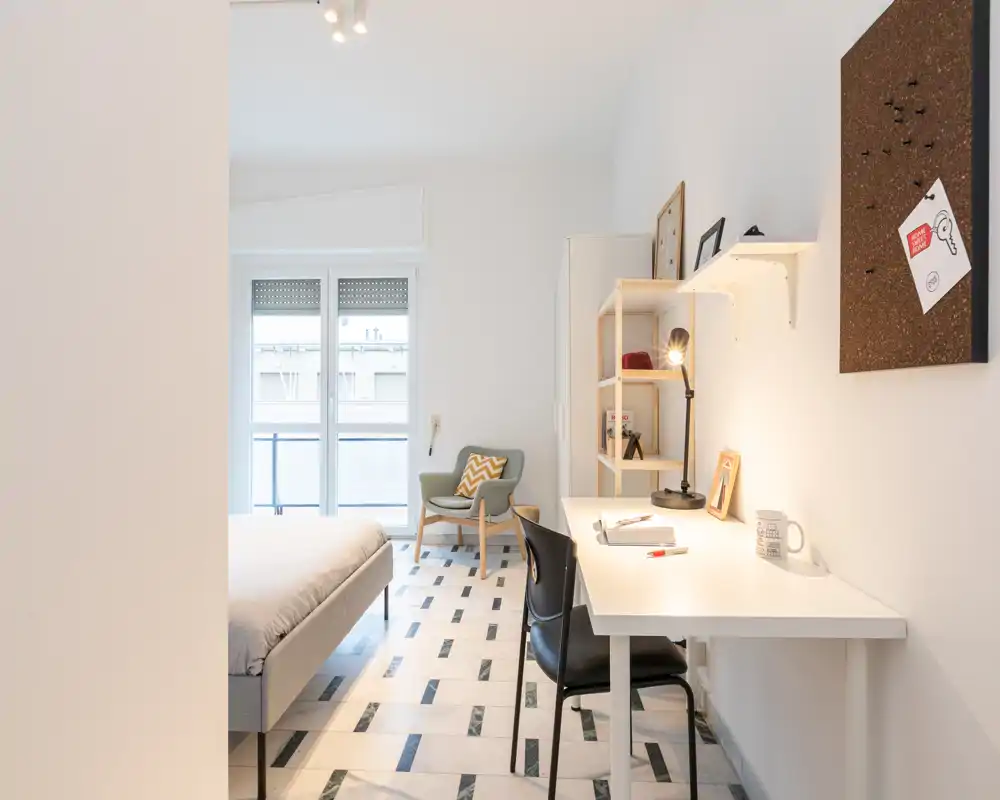 Bright, modern bedroom with a comfortable double bed, workspace with desk and lamp, and a cozy armchair by the window — ideal for both rest and study.