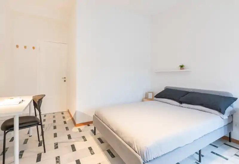 Bright, minimal bedroom with a fully visible double bed, simple bedside table and a small desk. The room feels clean and airy with neutral tones and natural light.