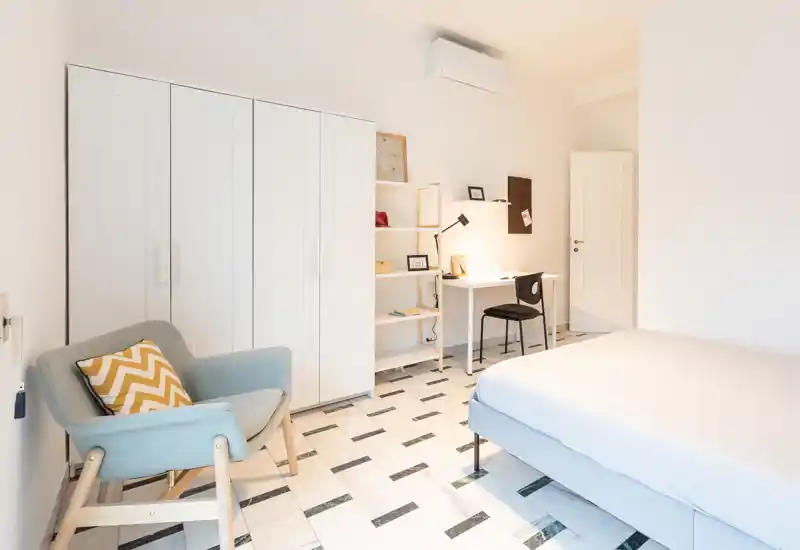 Bright, minimal bedroom with a comfortable double bed, a cozy armchair and a compact study desk; clean white wardrobes and patterned tile flooring give a fresh, airy feel.